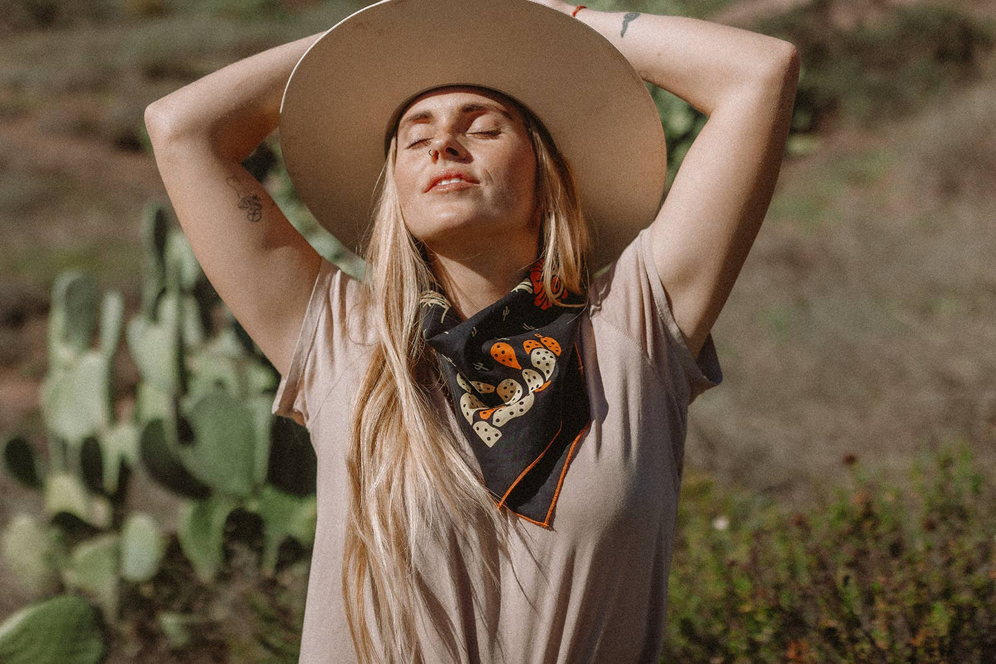 Model wearing the Bandits Cactus Dream bandana by Brothers Design Co. tied as a neckerchief while standing outdoors beside prickly pear cacti in warm sunlight.
