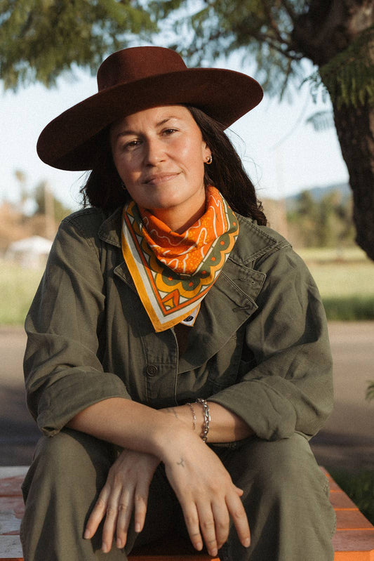 Model wearing the Bandits Sunset bandana by Studio MUTI tied as a neckerchief with an olive jacket and wide-brim hat in an outdoor setting.