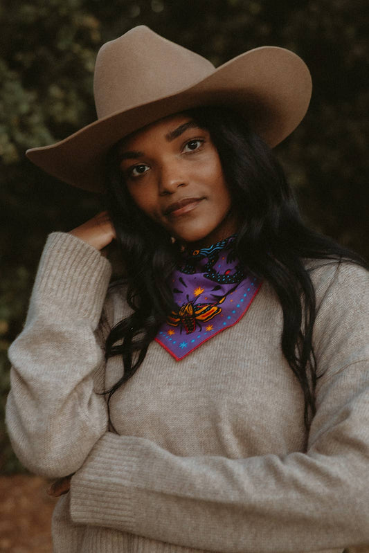 Model wearing the Pine Bones Electric Desert Parade bandana tied around the neck with a tan hat and cream sweater.