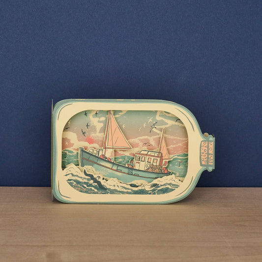 Tom Jay Fishing Boat in a Bottle die-cut greeting card featuring a fishing boat riding rolling waves beneath seabirds and a colorful sunset.