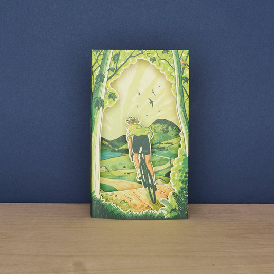 Front of the Tom Jay Spring Spins die-cut greeting card featuring a cyclist riding through a woodland trail toward rolling hills.
