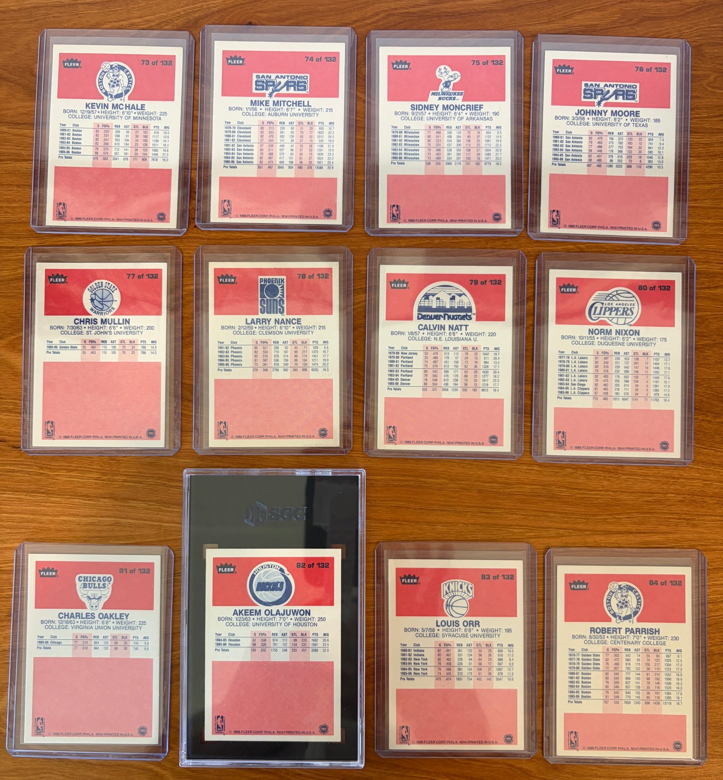 Backs of 1986 Fleer Basketball cards #73–84 from the complete 1986–87 Fleer Basketball set. Includes Kevin McHale #73, Sidney Moncrief #75, Chris Mullin #77, Larry Nance #78, Charles Oakley #81, Akeem Olajuwon #82 (SGC 7), Louis Orr #83, and Robert Parish #84. Card backs display player statistics, biographical information, and team logos. Cards are arranged in numerical order and shown in protective holders on a wood tabletop.