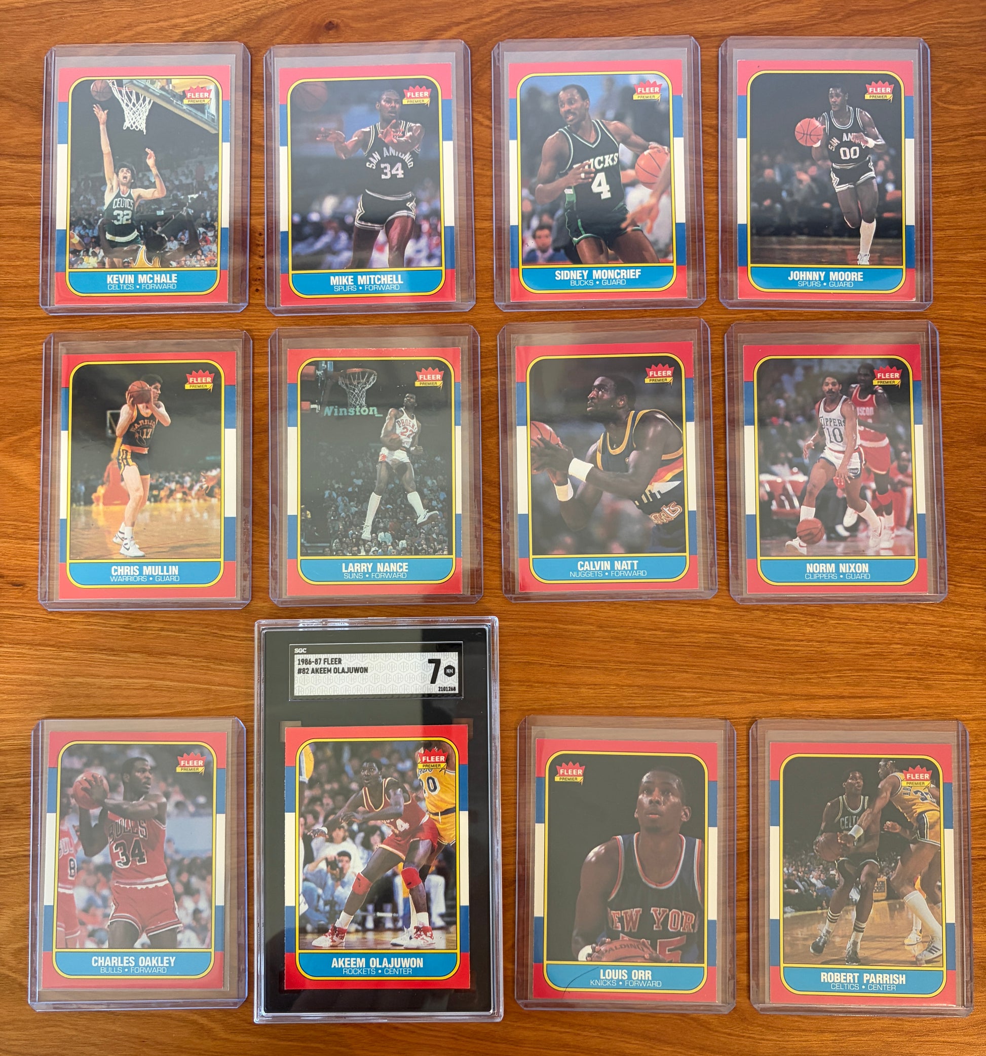 Fronts of 1986 Fleer Basketball cards #73–84 from the complete 1986–87 Fleer Basketball set. Includes Hall of Famers Akeem Olajuwon #82 (SGC 7), Kevin McHale #73, Chris Mullin #77, Charles Oakley #81, and Robert Parish #84, along with Mike Mitchell #74, Sidney Moncrief #75, Johnny Moore #76, Larry Nance #78, Calvin Natt #79, Norm Nixon #80, and Louis Orr #83. Cards are displayed in protective holders on a wood tabletop.