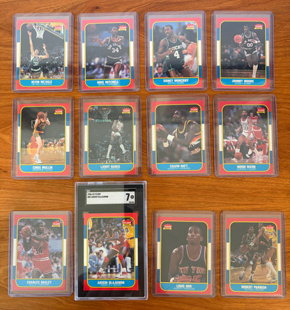 Fronts of 1986 Fleer Basketball cards #73–84 from the complete 1986–87 Fleer Basketball set. Includes Hall of Famers Akeem Olajuwon #82 (SGC 7), Kevin McHale #73, Chris Mullin #77, Charles Oakley #81, and Robert Parish #84, along with Mike Mitchell #74, Sidney Moncrief #75, Johnny Moore #76, Larry Nance #78, Calvin Natt #79, Norm Nixon #80, and Louis Orr #83. Cards are displayed in protective holders on a wood tabletop.