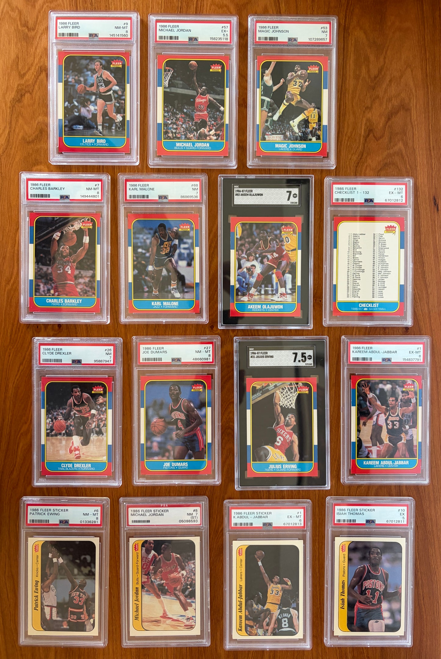Collection of graded key cards and stickers from the 1986 Fleer Basketball set displayed on a wood surface. Featured cards include Michael Jordan rookie card #57 PSA 5.5, Larry Bird #9 PSA 8, Magic Johnson #53 PSA 7, Charles Barkley rookie card #7 PSA 8, Karl Malone rookie card #68 PSA 7, Akeem Olajuwon rookie card #82 SGC 7, Clyde Drexler rookie card #26 PSA 7, Joe Dumars rookie card #26 PSA 8, Julius Erving #32 SGC 7.5, Kareem Abdul-Jabbar #1 PSA 6, and graded 1986 Fleer stickers including Michael Jordan 