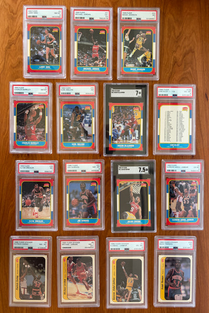 Collection of graded key cards and stickers from the 1986 Fleer Basketball set displayed on a wood surface. Featured cards include Michael Jordan rookie card #57 PSA 5.5, Larry Bird #9 PSA 8, Magic Johnson #53 PSA 7, Charles Barkley rookie card #7 PSA 8, Karl Malone rookie card #68 PSA 7, Akeem Olajuwon rookie card #82 SGC 7, Clyde Drexler rookie card #26 PSA 7, Joe Dumars rookie card #26 PSA 8, Julius Erving #32 SGC 7.5, Kareem Abdul-Jabbar #1 PSA 6, and graded 1986 Fleer stickers including Michael Jordan 