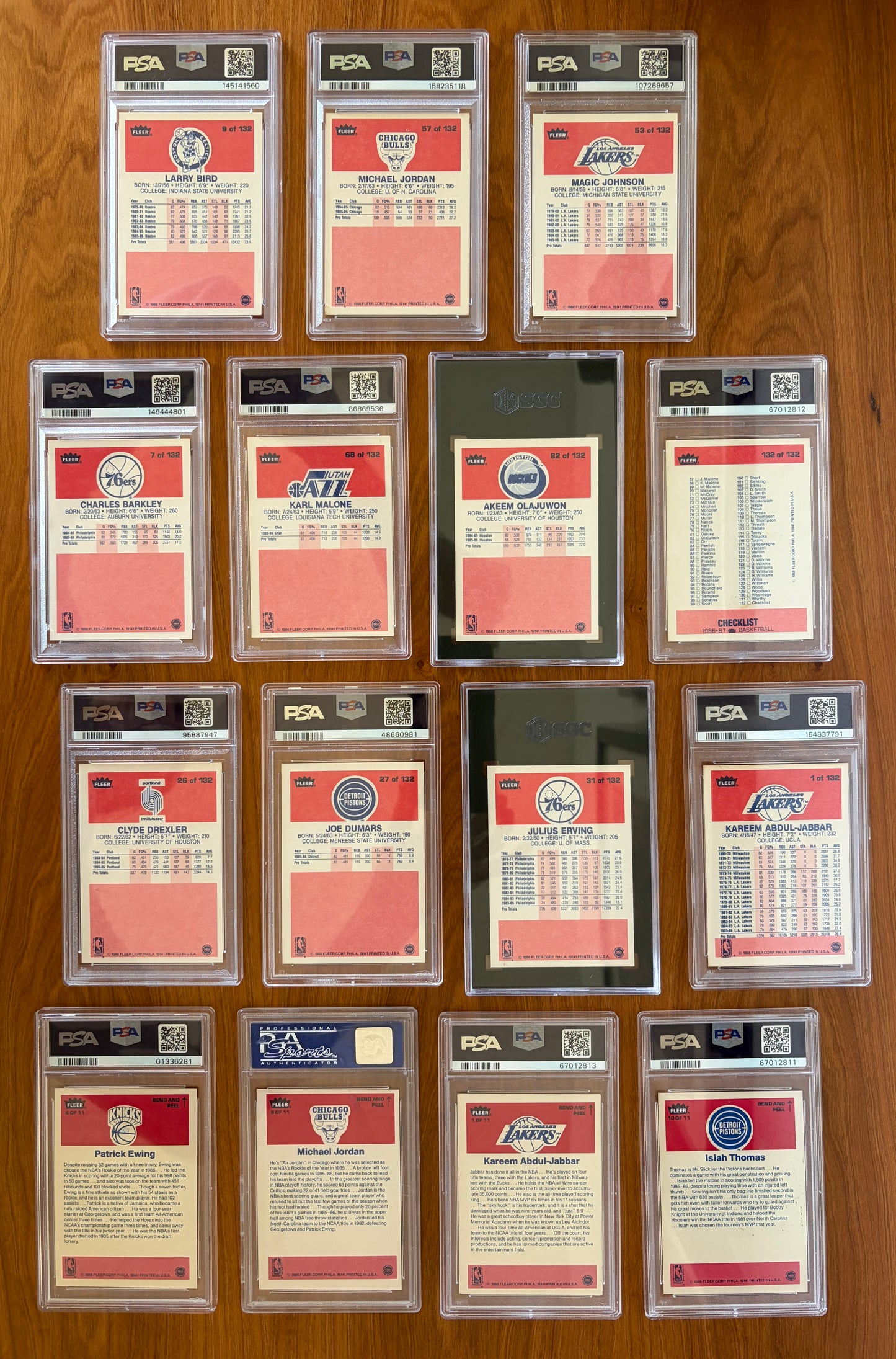 Backs of key graded cards and stickers from the 1986 Fleer Basketball set displayed on a wood surface. Featured cards include Michael Jordan rookie card #57 PSA 5.5, Larry Bird #9 PSA 8, Magic Johnson #53 PSA 7, Charles Barkley rookie card #7 PSA 8, Karl Malone rookie card #68 PSA 7, Hakeem Olajuwon rookie card #82 SGC 7, Clyde Drexler rookie card #26 PSA 7, Joe Dumars rookie card #27 PSA 8, Julius Erving #32 SGC 7.5, Kareem Abdul-Jabbar #1 PSA 6, and Checklist #132 PSA 6. Also shown are graded 1986 Fleer s