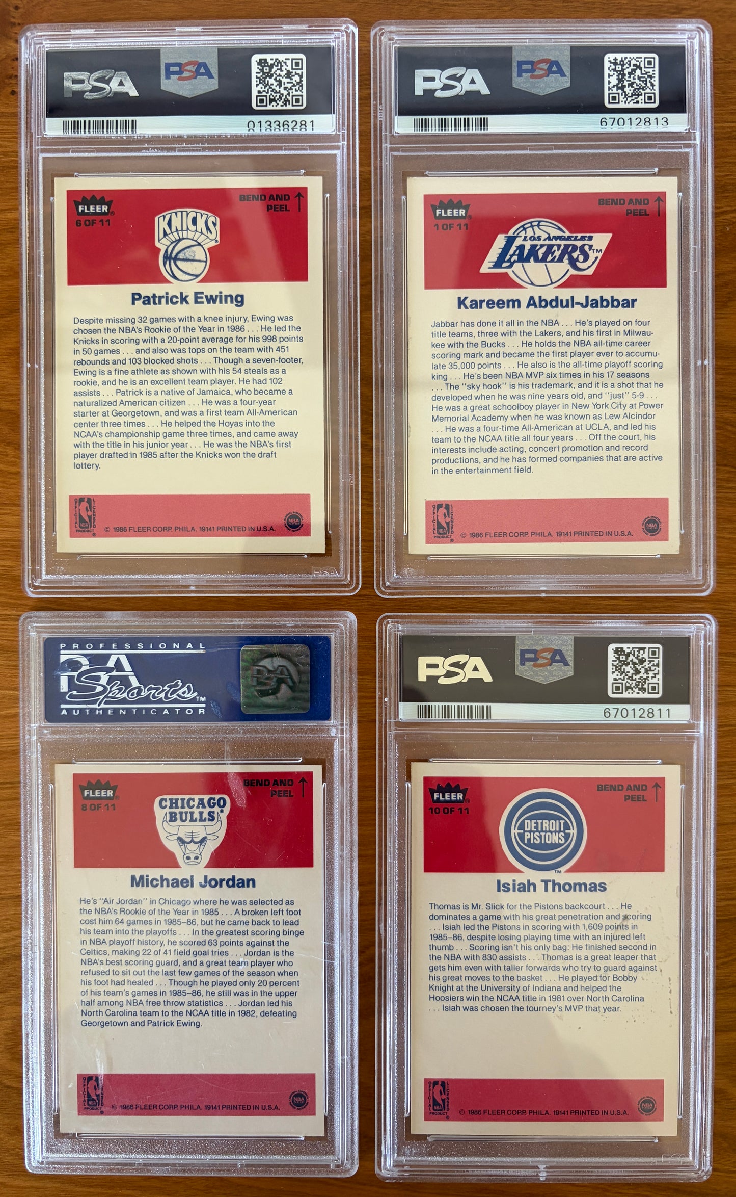 Backs of four graded 1986 Fleer Basketball stickers displayed on a wood surface. Featured stickers include Patrick Ewing #6 PSA 8, Kareem Abdul-Jabbar #1 PSA 6, Michael Jordan #8 PSA 7, and Isiah Thomas #10 PSA 5. The sticker backs contain player biographies and career highlights and are encapsulated in authenticated PSA holders.