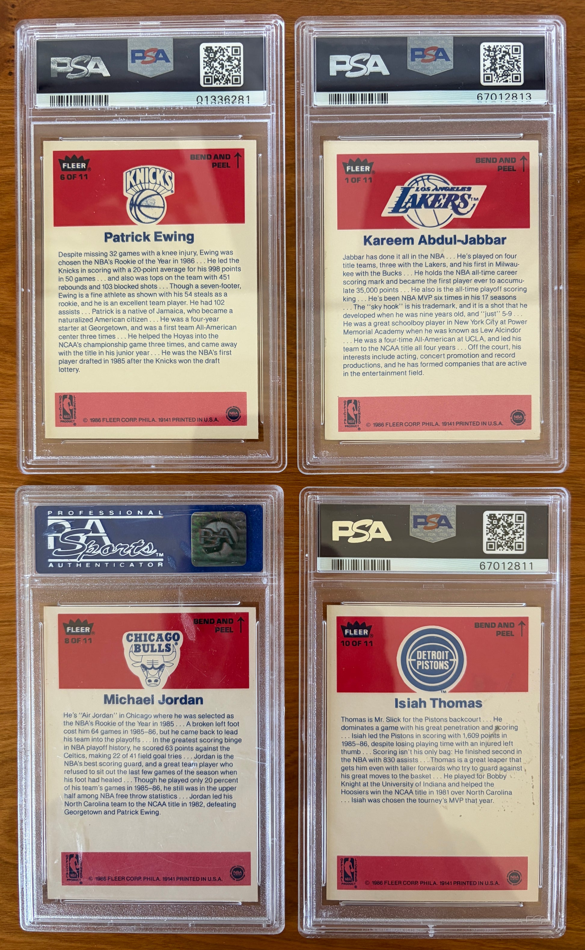 Backs of four graded 1986 Fleer Basketball stickers displayed on a wood surface. Featured stickers include Patrick Ewing #6 PSA 8, Kareem Abdul-Jabbar #1 PSA 6, Michael Jordan #8 PSA 7, and Isiah Thomas #10 PSA 5. The sticker backs contain player biographies and career highlights and are encapsulated in authenticated PSA holders.