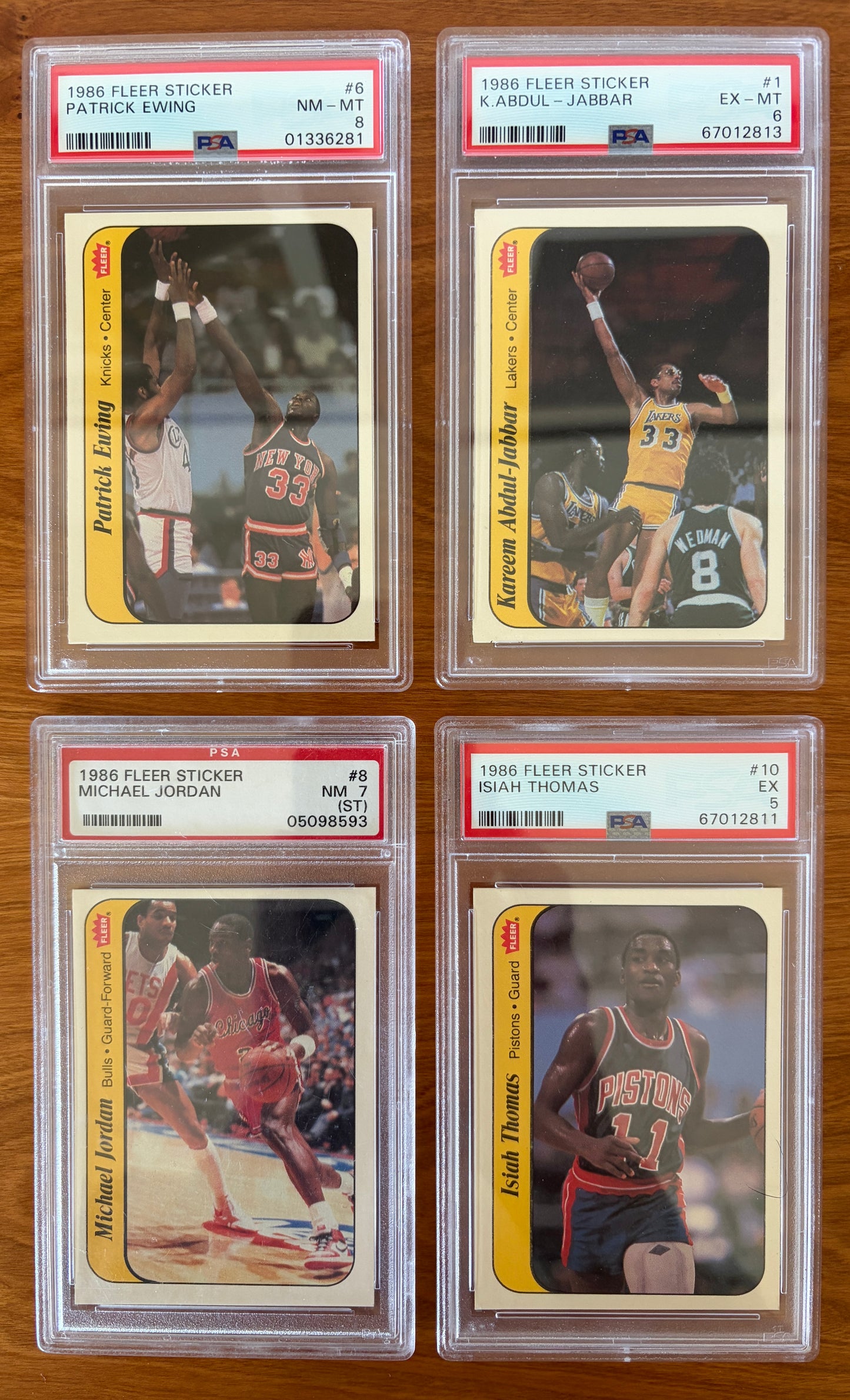 Four graded 1986 Fleer Basketball stickers displayed on a wood surface: Patrick Ewing sticker #6 PSA 8, Kareem Abdul-Jabbar sticker #1 PSA 6, Michael Jordan sticker #8 PSA 7, and Isiah Thomas sticker #10 PSA 5. The stickers feature Hall of Fame players from the iconic 1986 Fleer Basketball sticker set and are encapsulated in authenticated PSA holders.