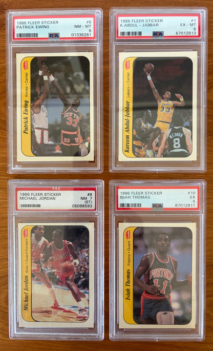 Four graded 1986 Fleer Basketball stickers displayed on a wood surface: Patrick Ewing sticker #6 PSA 8, Kareem Abdul-Jabbar sticker #1 PSA 6, Michael Jordan sticker #8 PSA 7, and Isiah Thomas sticker #10 PSA 5. The stickers feature Hall of Fame players from the iconic 1986 Fleer Basketball sticker set and are encapsulated in authenticated PSA holders.