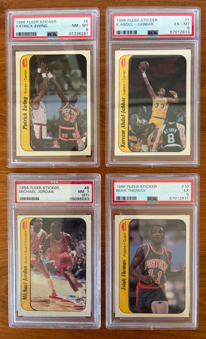 Four graded 1986 Fleer Basketball stickers displayed on a wood surface: Patrick Ewing sticker #6 PSA 8, Kareem Abdul-Jabbar sticker #1 PSA 6, Michael Jordan sticker #8 PSA 7, and Isiah Thomas sticker #10 PSA 5. The stickers feature Hall of Fame players from the iconic 1986 Fleer Basketball sticker set and are encapsulated in authenticated PSA holders.