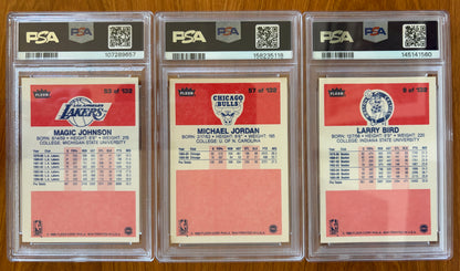 Backs of three graded 1986 Fleer basketball cards displayed side by side: Magic Johnson #53 PSA 7, Michael Jordan rookie card #57 PSA 5.5, and Larry Bird #9 PSA 8. The card backs show player biographies, career statistics, and team logos for the Los Angeles Lakers, Chicago Bulls, and Boston Celtics.
