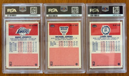 Backs of three graded 1986 Fleer basketball cards displayed side by side: Magic Johnson #53 PSA 7, Michael Jordan rookie card #57 PSA 5.5, and Larry Bird #9 PSA 8. The card backs show player biographies, career statistics, and team logos for the Los Angeles Lakers, Chicago Bulls, and Boston Celtics.