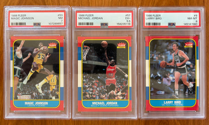 Three graded 1986 Fleer basketball cards displayed side by side: Magic Johnson #53 PSA 7, Michael Jordan rookie card #57 PSA 5.5, and Larry Bird #9 PSA 8. The cards feature three of the most iconic players in basketball history and are part of the complete 1986 Fleer Basketball set.