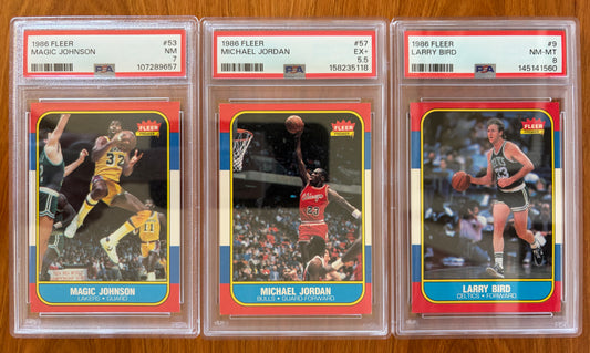 Three graded 1986 Fleer basketball cards displayed side by side: Magic Johnson #53 PSA 7, Michael Jordan rookie card #57 PSA 5.5, and Larry Bird #9 PSA 8. The cards feature three of the most iconic players in basketball history and are part of the complete 1986 Fleer Basketball set.