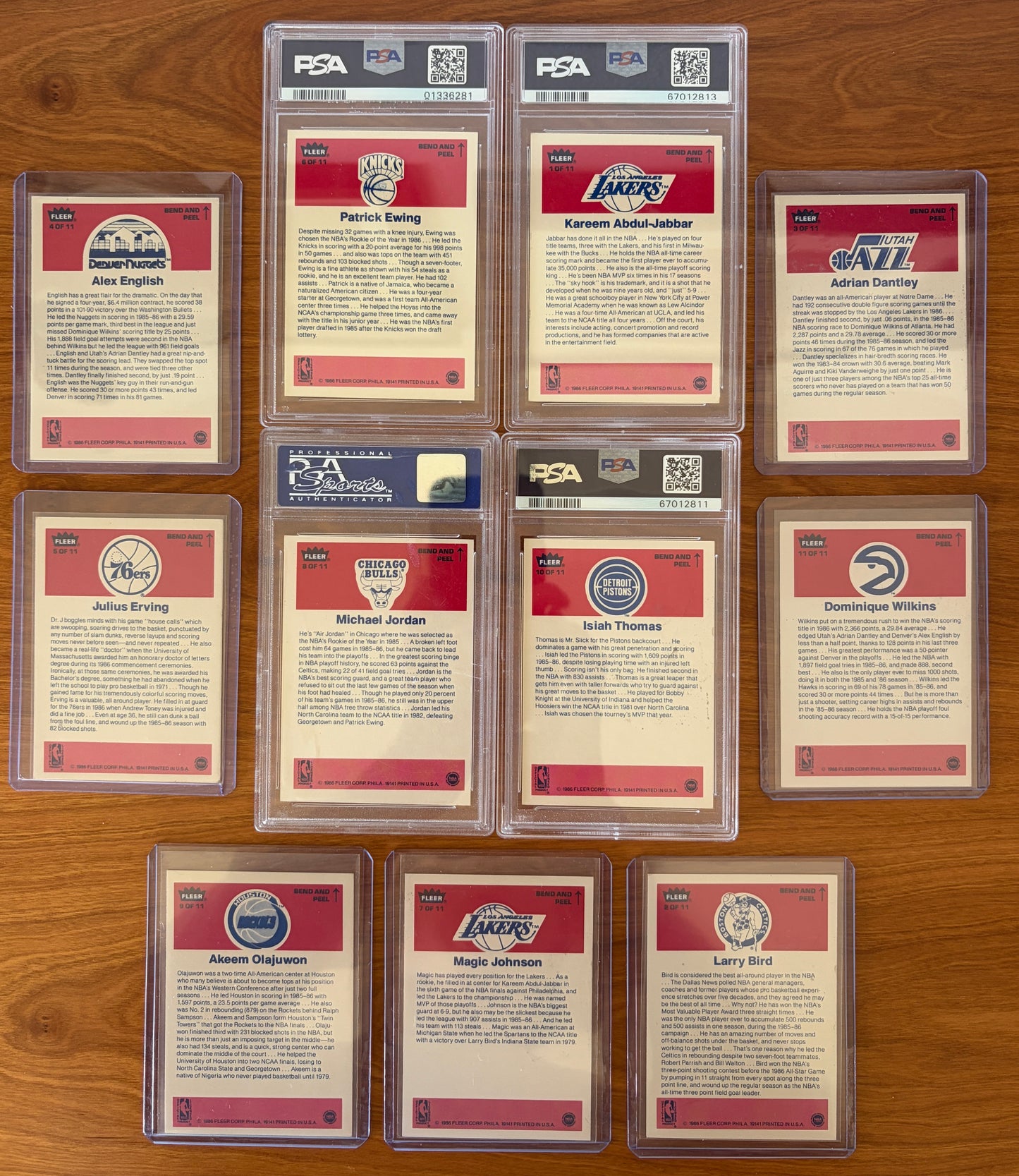 Backs of the 1986 Fleer Basketball sticker set displayed on a wood surface. Featured are graded stickers of Patrick Ewing PSA 8, Kareem Abdul-Jabbar PSA 6, Michael Jordan PSA 7, and Isiah Thomas PSA 5, alongside ungraded stickers of Alex English, Adrian Dantley, Julius Erving, Dominique Wilkins, Hakeem Olajuwon, Magic Johnson, and Larry Bird. The sticker backs include player biographies, career highlights, and team logos from the iconic 1986 Fleer Basketball release.