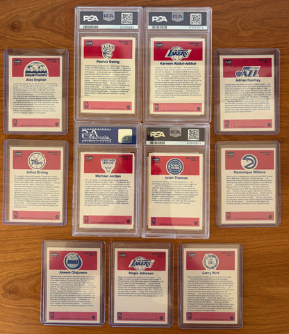 Backs of the 1986 Fleer Basketball sticker set displayed on a wood surface. Featured are graded stickers of Patrick Ewing PSA 8, Kareem Abdul-Jabbar PSA 6, Michael Jordan PSA 7, and Isiah Thomas PSA 5, alongside ungraded stickers of Alex English, Adrian Dantley, Julius Erving, Dominique Wilkins, Hakeem Olajuwon, Magic Johnson, and Larry Bird. The sticker backs include player biographies, career highlights, and team logos from the iconic 1986 Fleer Basketball release.