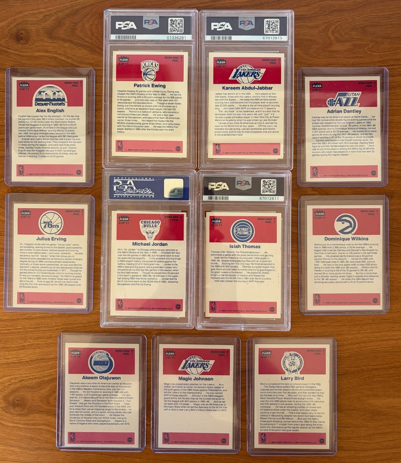 Backs of the 1986 Fleer Basketball sticker set displayed on a wood surface. Featured are graded stickers of Patrick Ewing PSA 8, Kareem Abdul-Jabbar PSA 6, Michael Jordan PSA 7, and Isiah Thomas PSA 5, alongside ungraded stickers of Alex English, Adrian Dantley, Julius Erving, Dominique Wilkins, Hakeem Olajuwon, Magic Johnson, and Larry Bird. The sticker backs include player biographies, career highlights, and team logos from the iconic 1986 Fleer Basketball release.