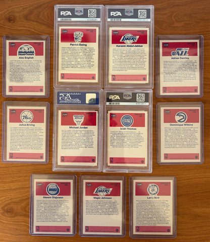 Backs of the 1986 Fleer Basketball sticker set displayed on a wood surface. Featured are graded stickers of Patrick Ewing PSA 8, Kareem Abdul-Jabbar PSA 6, Michael Jordan PSA 7, and Isiah Thomas PSA 5, alongside ungraded stickers of Alex English, Adrian Dantley, Julius Erving, Dominique Wilkins, Hakeem Olajuwon, Magic Johnson, and Larry Bird. The sticker backs include player biographies, career highlights, and team logos from the iconic 1986 Fleer Basketball release.