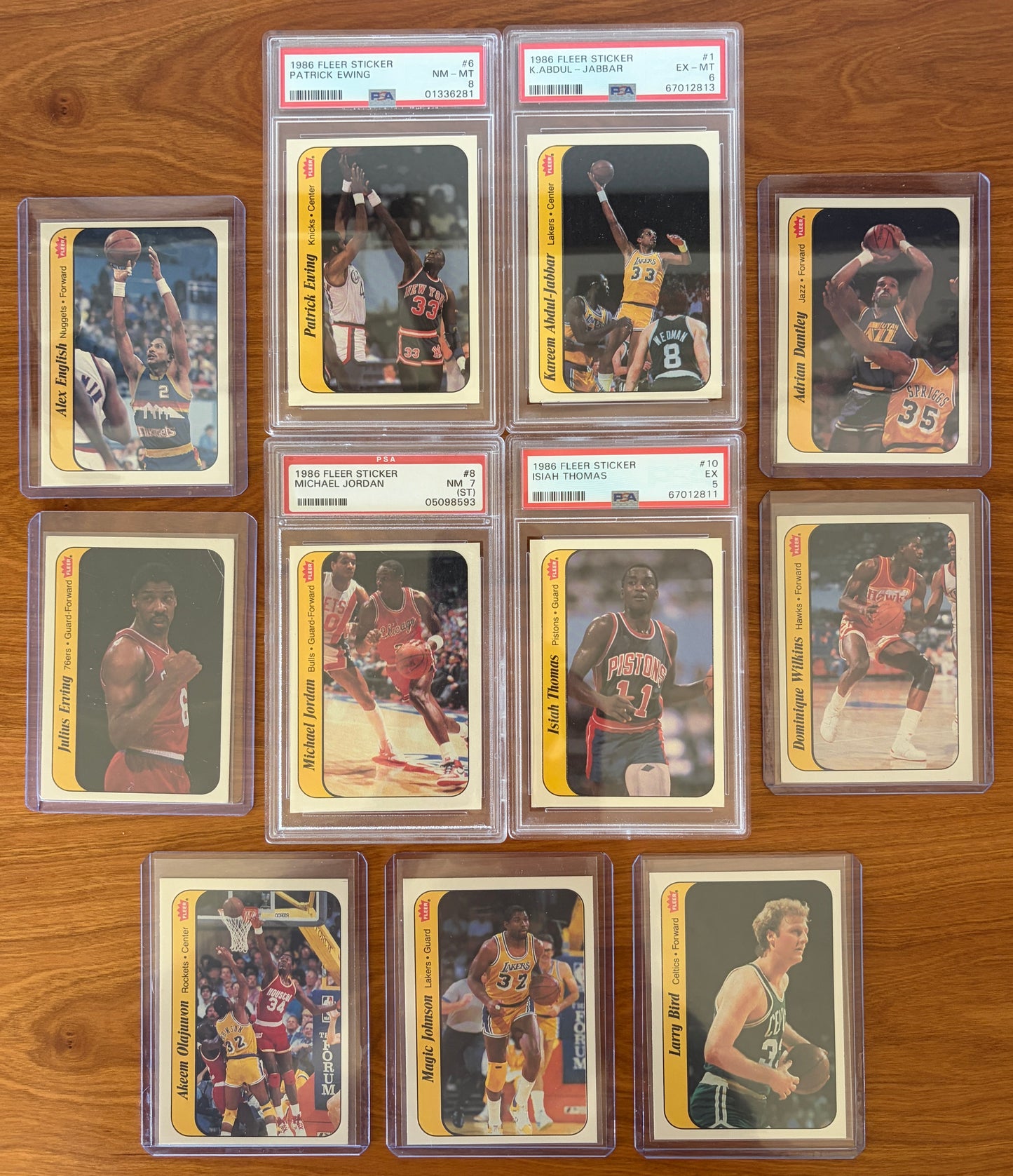 1986 Fleer Basketball sticker set displayed on a wood surface featuring graded stickers of Michael Jordan PSA 7, Patrick Ewing PSA 8, Kareem Abdul-Jabbar PSA 6, and Isiah Thomas PSA 5, alongside ungraded stickers of Larry Bird, Magic Johnson, Hakeem Olajuwon, Julius Erving, Dominique Wilkins, Adrian Dantley, and Alex English. The iconic sticker set showcases many of basketball's greatest Hall of Fame players from the legendary 1986 Fleer release.