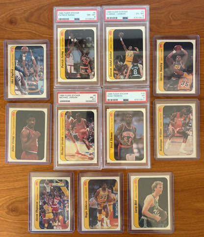 1986 Fleer Basketball sticker set displayed on a wood surface featuring graded stickers of Michael Jordan PSA 7, Patrick Ewing PSA 8, Kareem Abdul-Jabbar PSA 6, and Isiah Thomas PSA 5, alongside ungraded stickers of Larry Bird, Magic Johnson, Hakeem Olajuwon, Julius Erving, Dominique Wilkins, Adrian Dantley, and Alex English. The iconic sticker set showcases many of basketball's greatest Hall of Fame players from the legendary 1986 Fleer release.