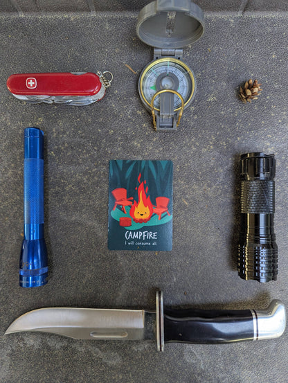 Collection of outdoor survival items including a knife, Swiss Army knife, compass, flashlights, and a card with a campfire illustration on a gray surface.