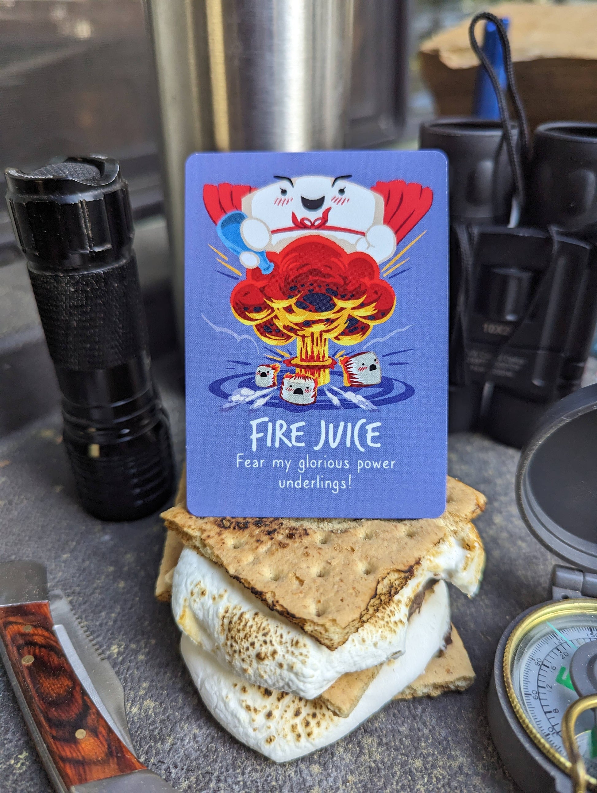 Card with cartoon character and text 'Fire Juice' on a s'mores stack with a knife, compass, and pepper grinder in the background.