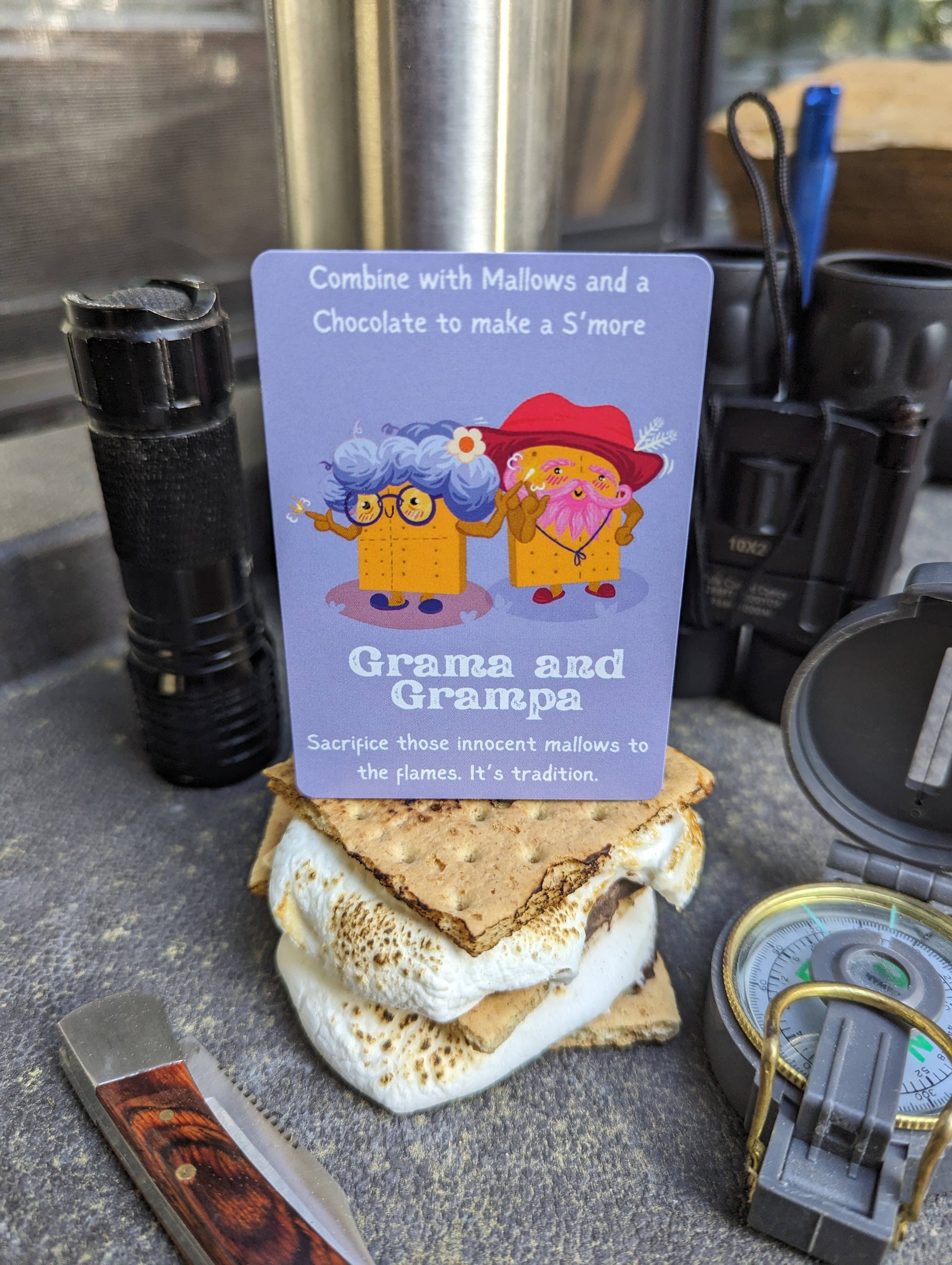 S'mores card with cartoon characters on a camping scene with flashlight, knife, and compass.