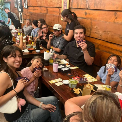 Group of people sitting around a wooden table in a casual dining setting, enjoying drinks and food while playing Roast