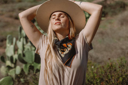 Model wearing the Bandits Cactus Dream bandana by Brothers Design Co. tied as a neckerchief while standing outdoors beside prickly pear cacti in warm sunlight.