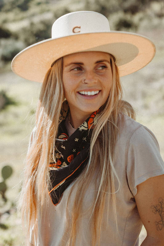 Model wearing the Bandits Cactus Dream bandana by Brothers Design Co. tied as a neck scarf with a wide-brim hat in a desert setting.