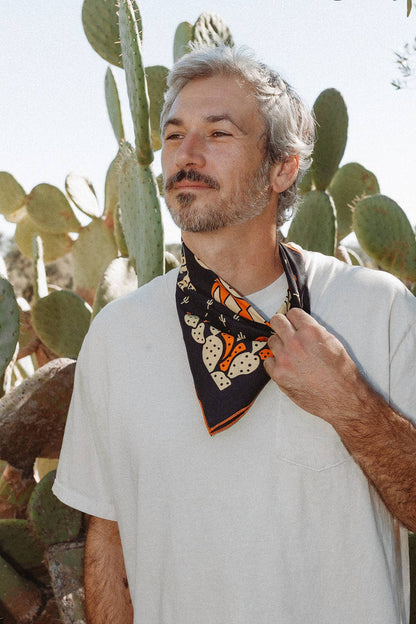 Model wearing the Bandits Cactus Dream bandana by Brothers Design Co. tied as a neckerchief with a white T-shirt in front of prickly pear cacti.