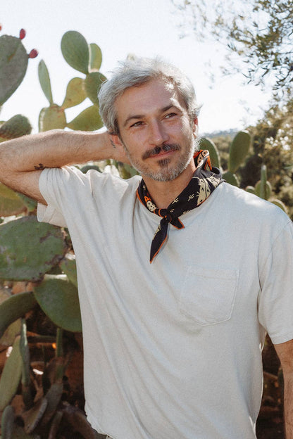 Model wearing the Bandits Cactus Dream bandana by Brothers Design Co. tied as a neckerchief with a white T-shirt while standing among prickly pear cacti.