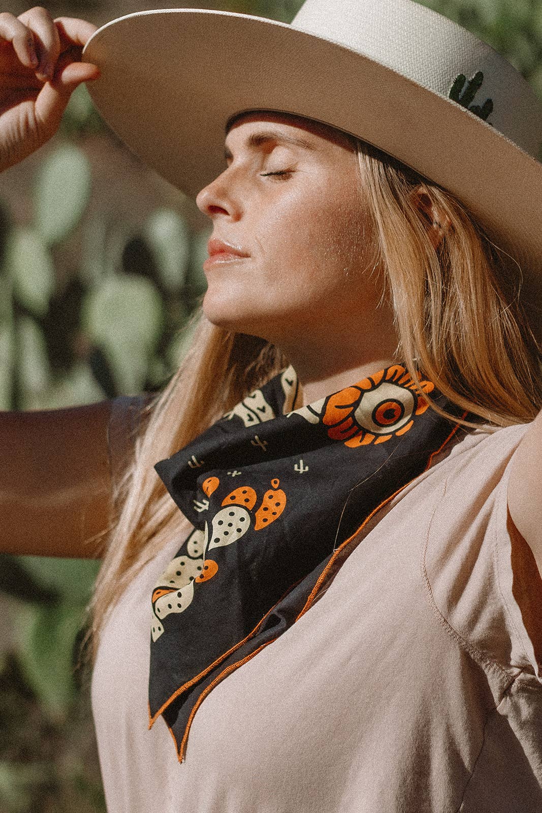 Model wearing the Bandits Cactus Dream bandana by Brothers Design Co. tied as a neckerchief with a wide-brim hat in a desert setting.