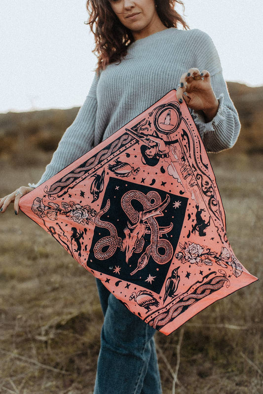 Model holding the Bandits Rattle & Hiss bandana by Nuri Amanatullah fully unfolded, showcasing the complete Southwestern-inspired artwork with a rattlesnake, longhorn skull, desert birds, and wildflowers.