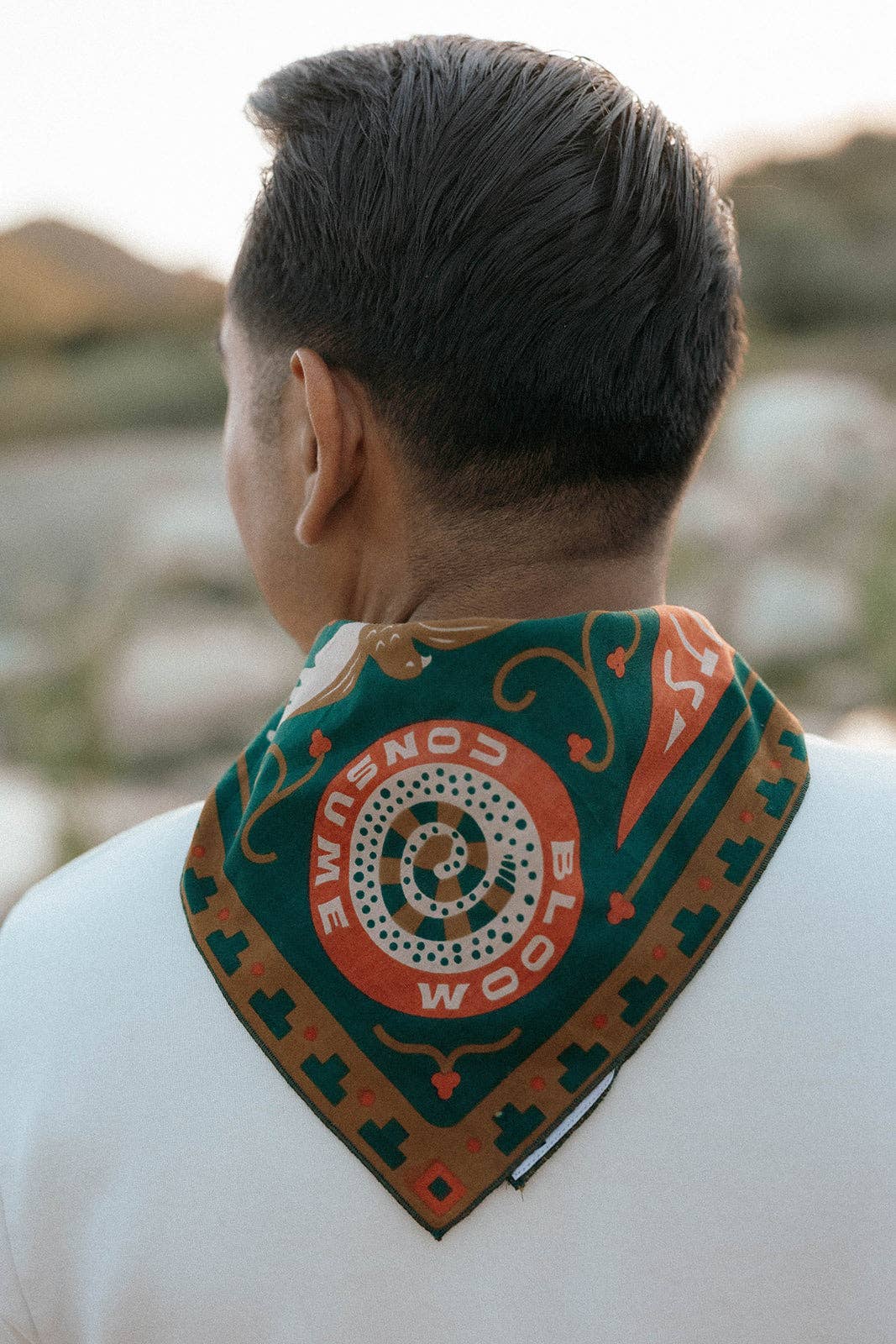 Rear view of a model wearing the Bandits Greenbelt bandana by Sam Larson around the neck, showing the "Bloom & Consume" emblem and nature-inspired artwork.