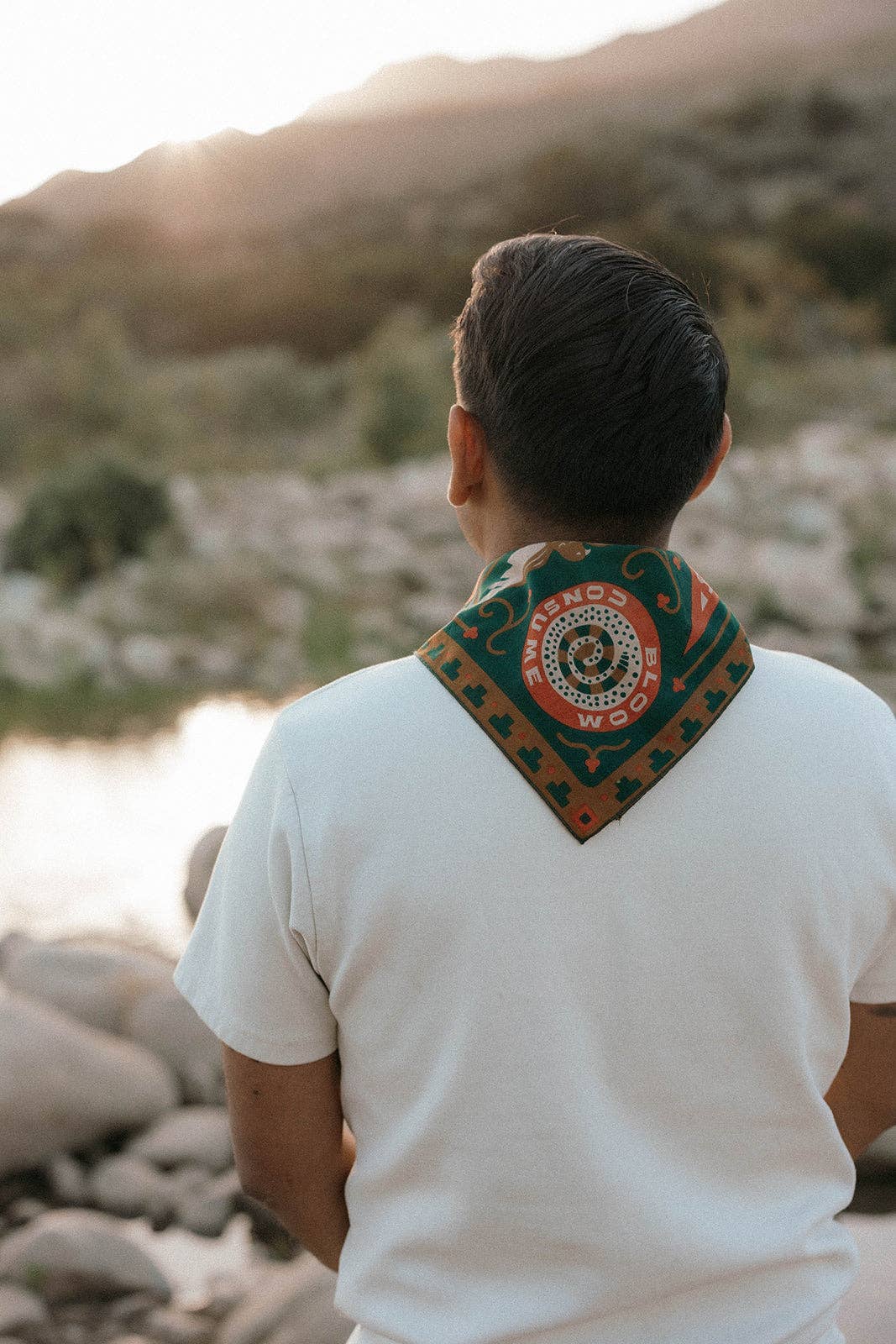 Rear view of a model wearing the Bandits Greenbelt bandana by Sam Larson around the neck beside a river, showing the circular "Bloom & Consume" emblem and nature-inspired artwork.
