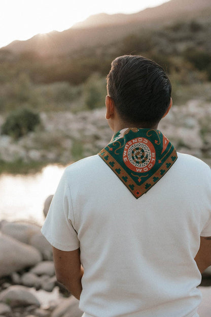 Rear view of a model wearing the Bandits Greenbelt bandana by Sam Larson around the neck beside a river, showing the circular "Bloom & Consume" emblem and nature-inspired artwork.