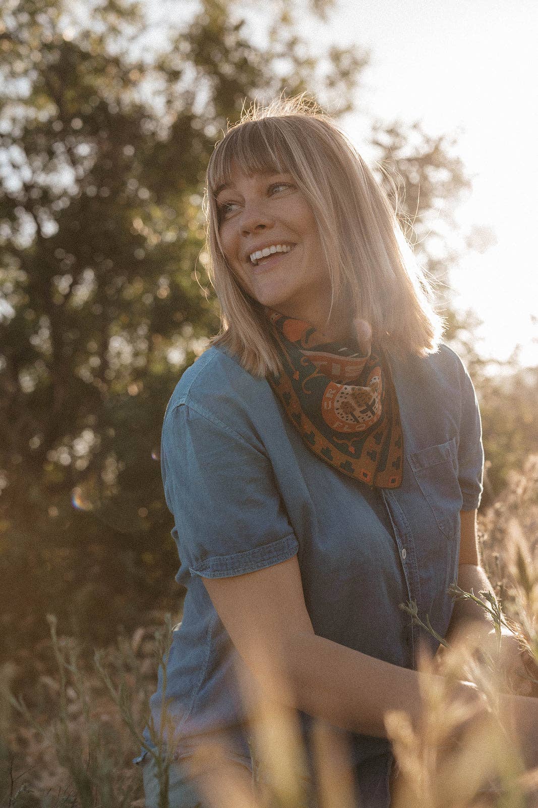 Model wearing the Bandits Greenbelt bandana by Sam Larson tied as a neckerchief while sitting outdoors in warm evening light.
