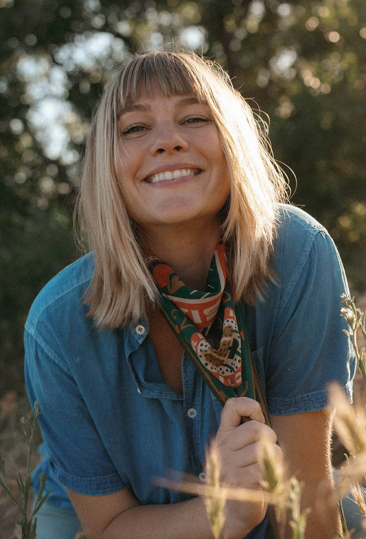 Model wearing the Bandits Greenbelt bandana by Sam Larson tied as a neck scarf with a blue button-up shirt in an outdoor setting.