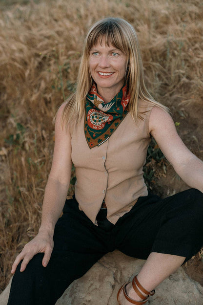 Model wearing the Bandits Greenbelt bandana by Sam Larson tied as a neckerchief with a sleeveless tan top while seated outdoors.