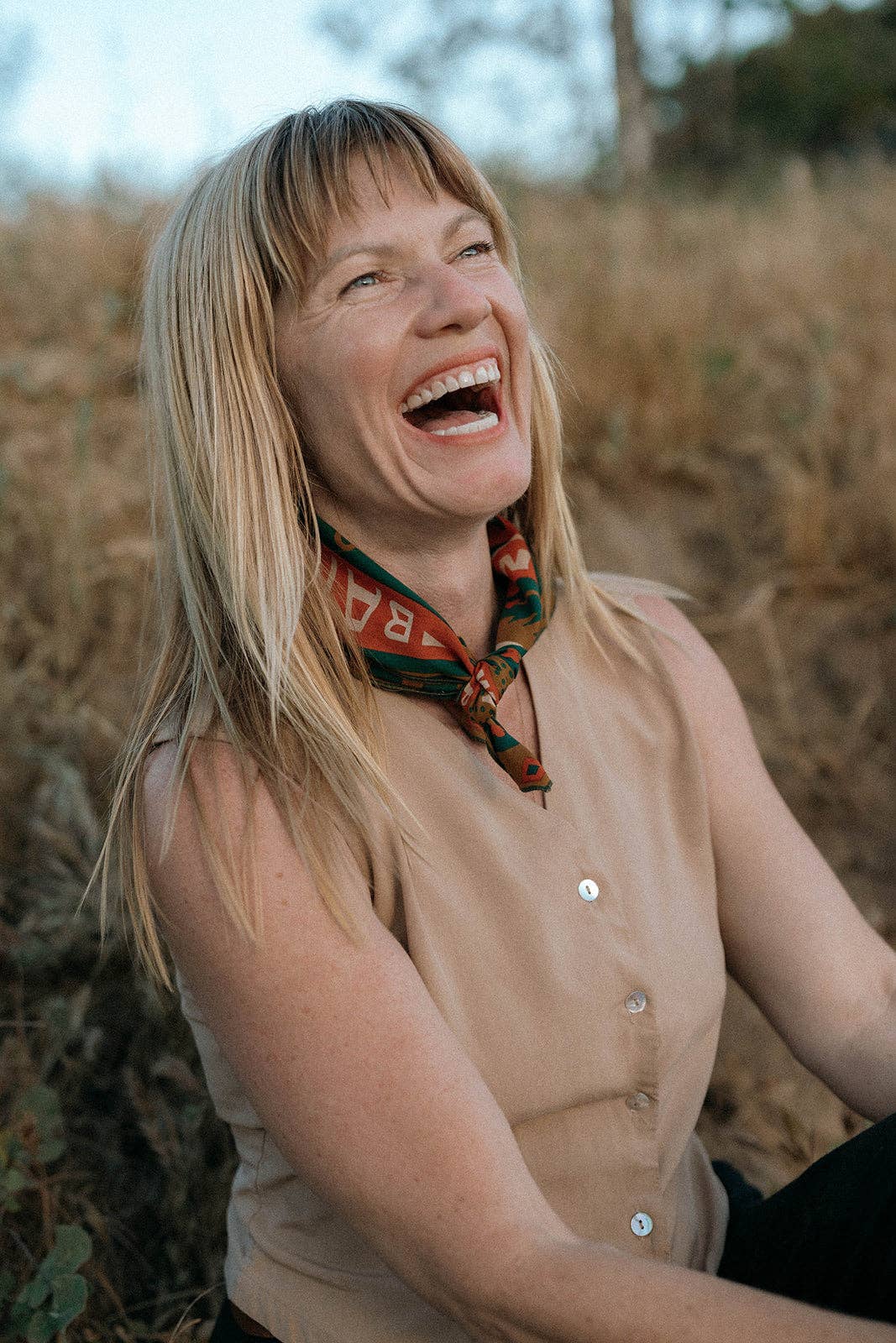 Model wearing the Bandits Greenbelt bandana by Sam Larson tied as a neckerchief with a sleeveless tan top in an outdoor setting.