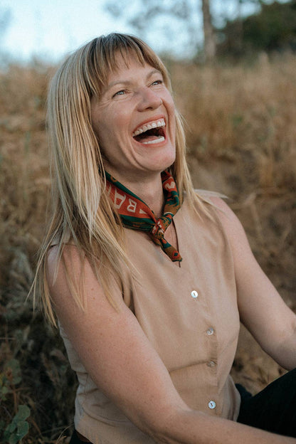 Model wearing the Bandits Greenbelt bandana by Sam Larson tied as a neckerchief with a sleeveless tan top in an outdoor setting.