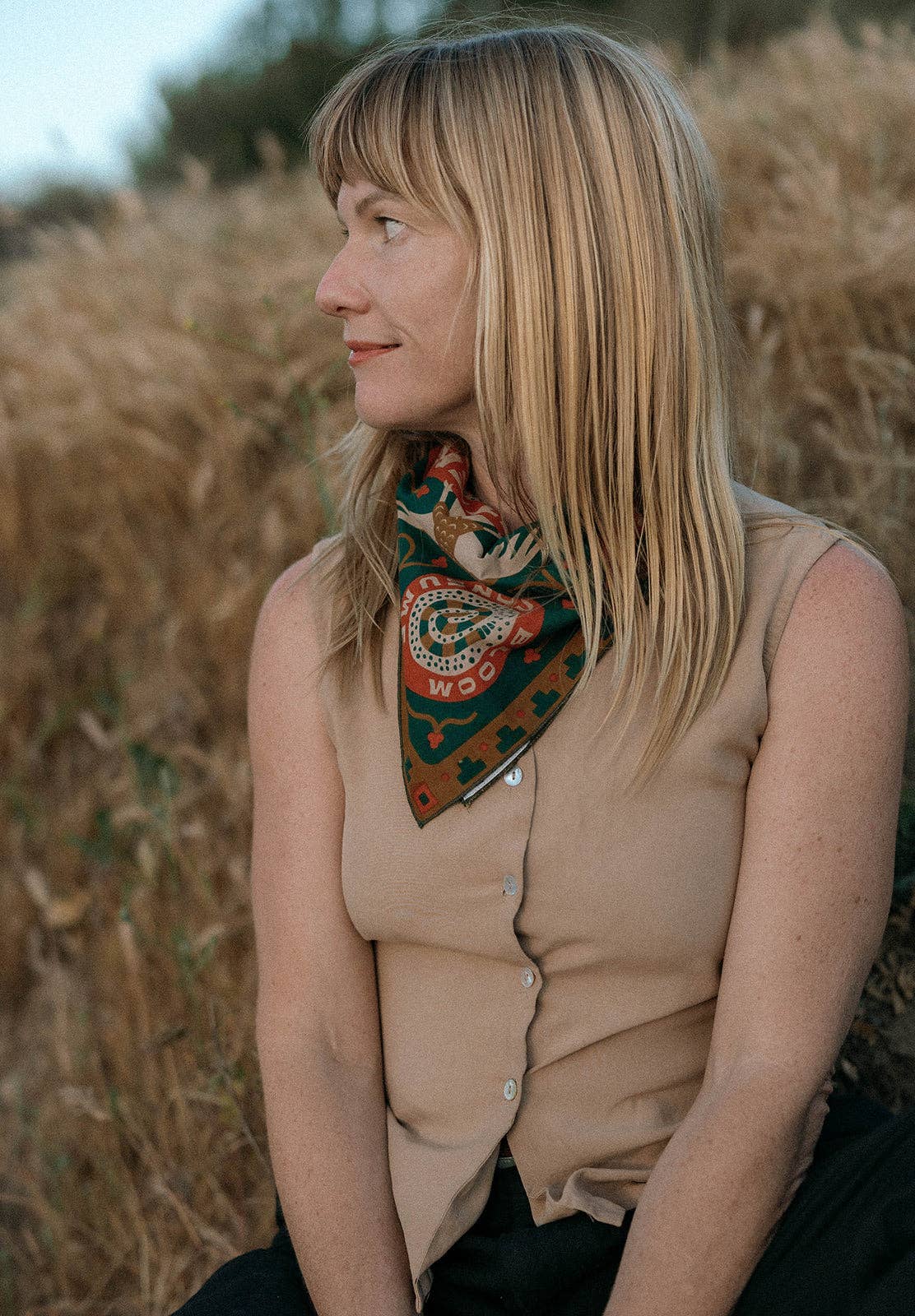 Model wearing the Bandits Greenbelt bandana by Sam Larson tied as a neckerchief with a sleeveless tan top, showing the nature-inspired artwork and circular "Bloom & Consume" emblem.