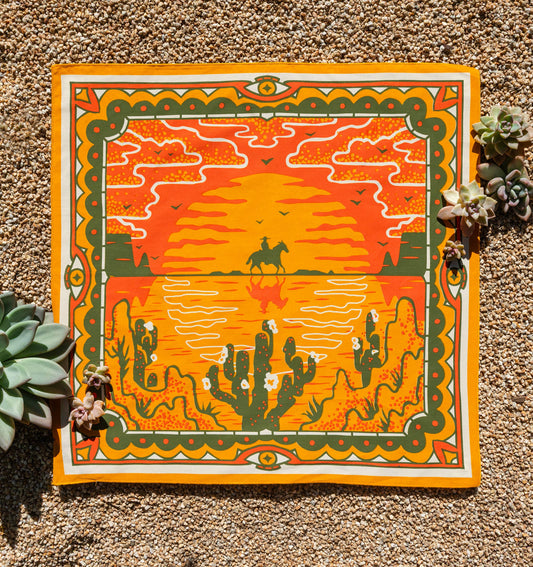 Bandits Sunset bandana by Studio MUTI featuring a rider on horseback silhouetted against a glowing sunset, with desert cacti, birds, and warm landscape-inspired artwork.
