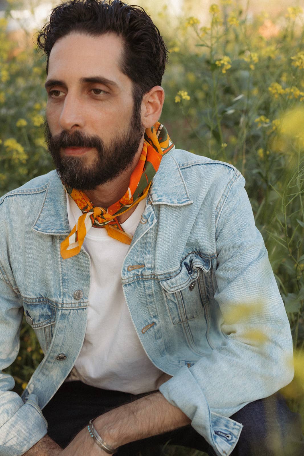 Model wearing the Bandits Sunset bandana by Studio MUTI tied as a rolled neck scarf with a denim jacket in a field of wildflowers.
