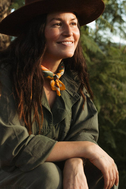 Model wearing the Bandits Sunset bandana by Studio MUTI tied as a neck tie with an olive jacket and wide-brim hat in warm evening light.