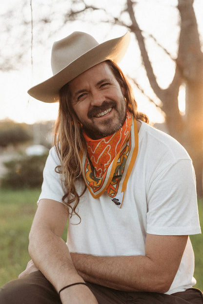 Model wearing the Bandits Sunset bandana by Studio MUTI tied as a neckerchief with a white T-shirt and wide-brim hat in warm evening light.