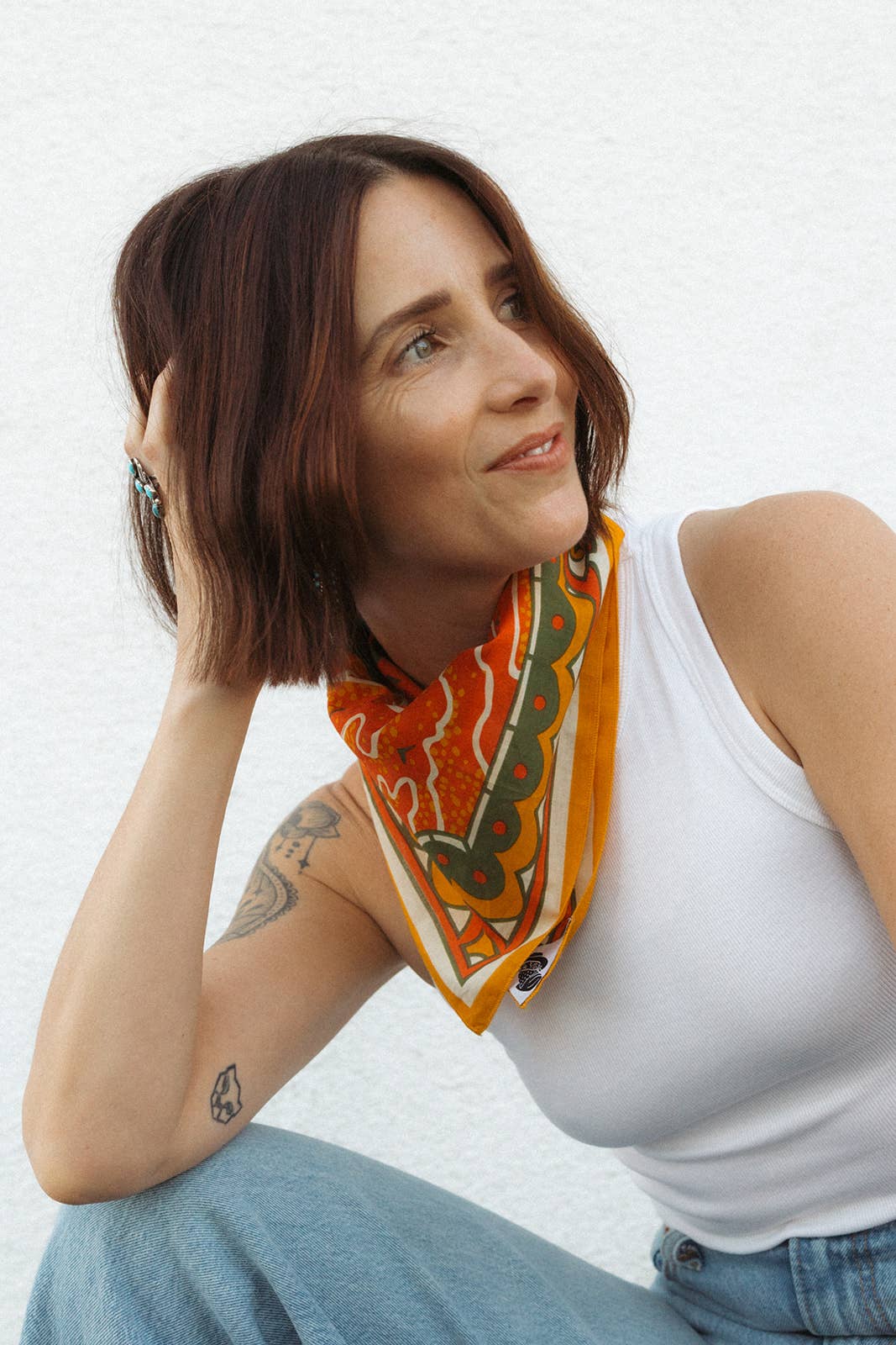 Model wearing the Bandits Sunset bandana by Studio MUTI tied as a neckerchief with a white tank top, showcasing the warm orange and olive landscape-inspired artwork.